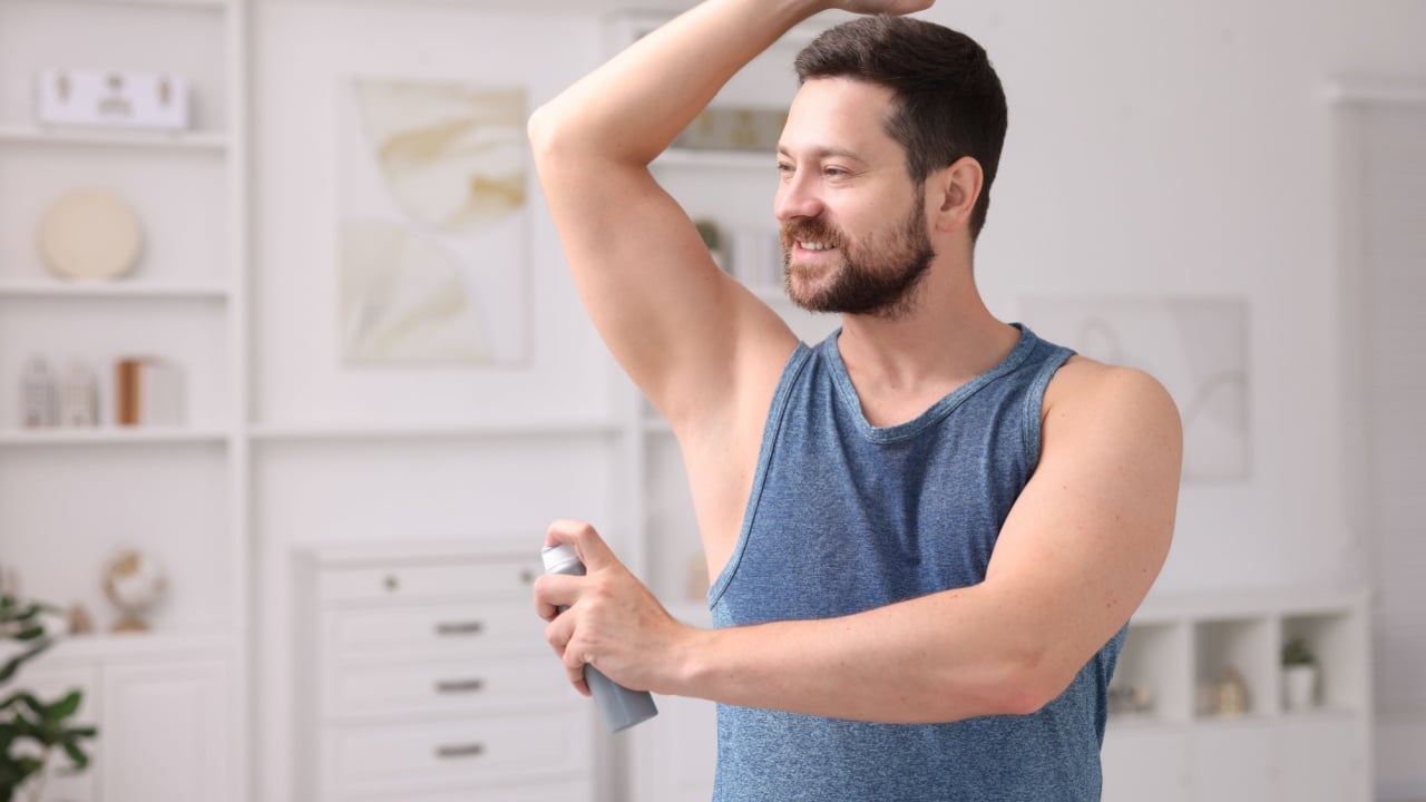 Smiling man applying spray deodorant at home. Space for text