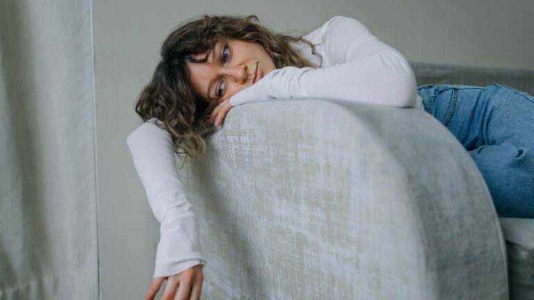 Young woman in casual clothes lounges on a sofa, lost in thought at home. The serene atmosphere adds to her loneliness, as natural light creates a cozy ambiance for her weekend daydreaming