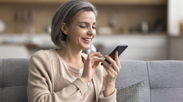 Home comfort and digital tech. Relaxed happy mature woman spend leisure time interacting with phone app on couch at living room text message to friend read electronic book browse websites.