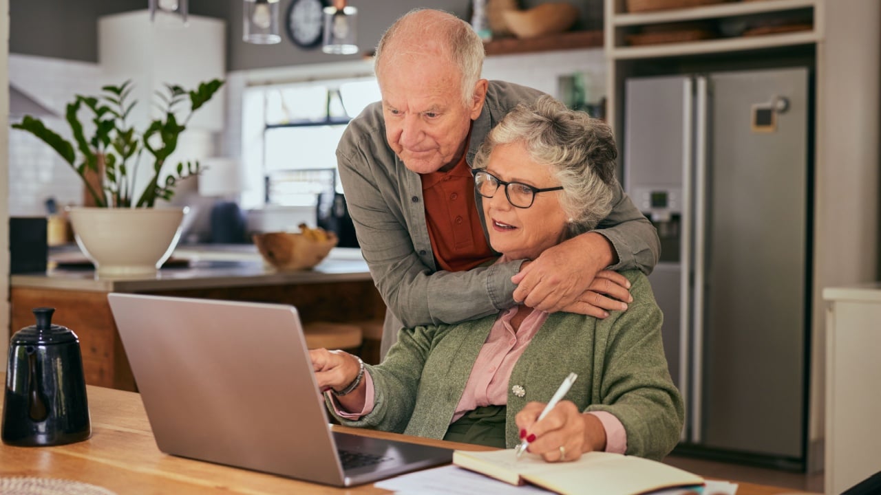 Elderly couple collaborating on tasks with a laptop and notebook at home while sharing an affectionate moment. Old man and elderly woman managing documents and finances together. Mature woman working.