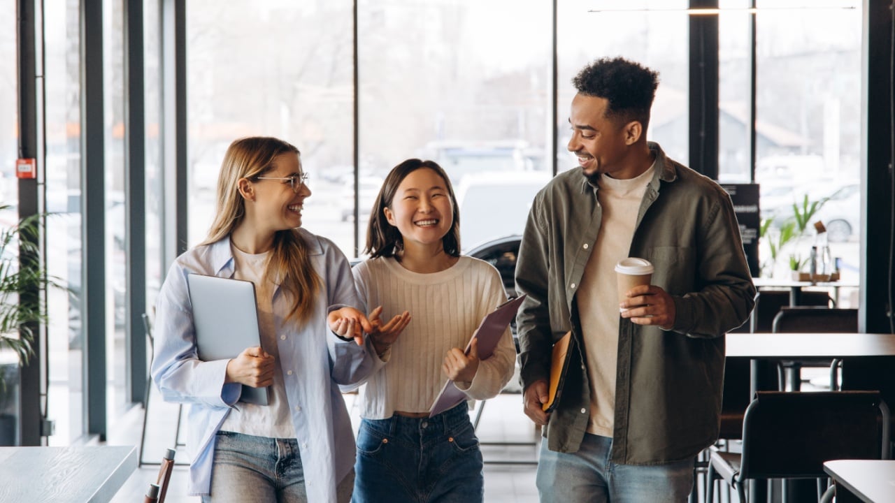Three cheerful multi-ethnic university students are walking through the university cafeteria, holding laptops and notebooks, engaging in friendly conversation and enjoying a coffee break