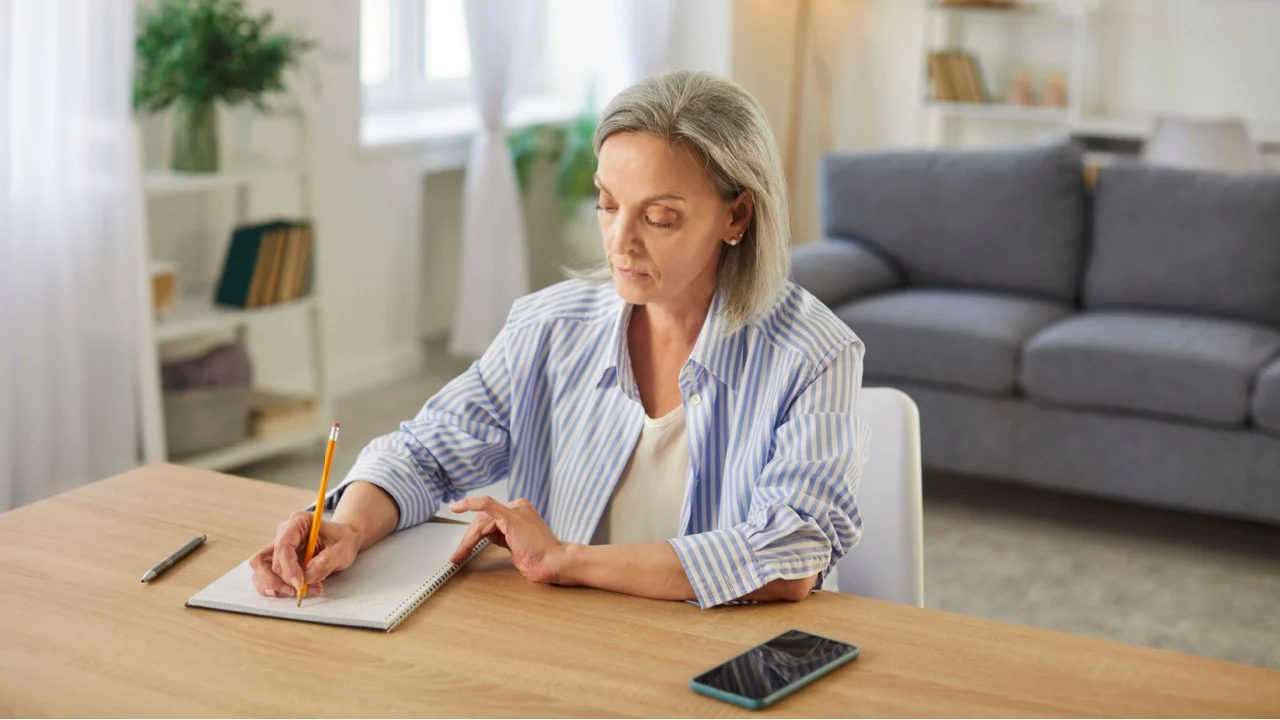 Retired old senior woman, pleasant elderly person alone at home, female sitting writing at desk, grandmother at home, handwriting letter making notes on paper, focused thinking in room calculating