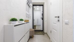 A clean and modern hallway featuring white walls, a mirror, a storage cabinet, a small ottoman, and decorative plants.