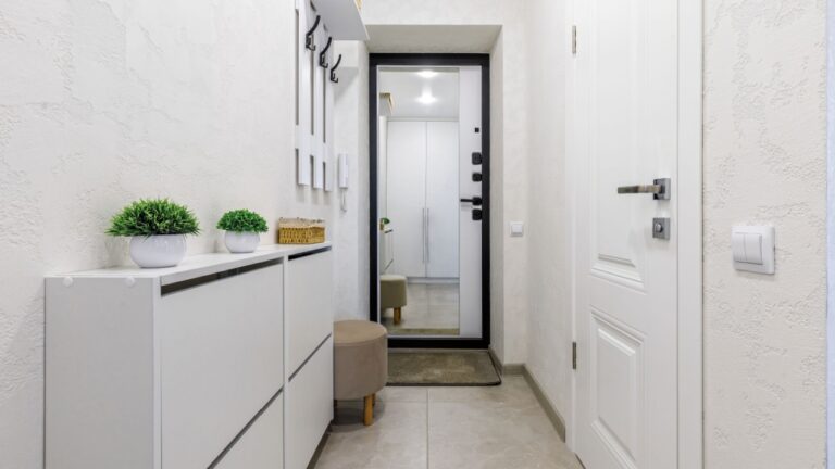A clean and modern hallway featuring white walls, a mirror, a storage cabinet, a small ottoman, and decorative plants.