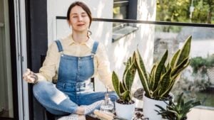 A young woman sits cross-legged, meditating with potted snake plants on a sunny balcony. A peaceful mix of mindfulness and urban gardening.
