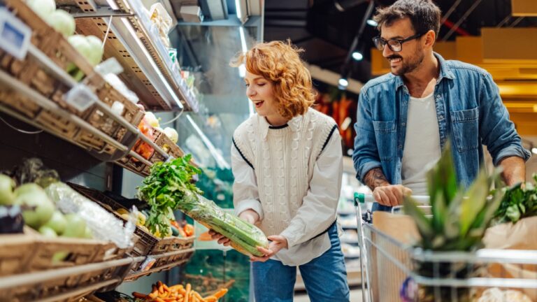 Young smiling woman picking celery while grocery shopping with her boyfriend, pushing a shopping cart full of fresh vegetables and other groceries