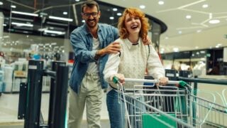 Cheerful man and woman are exiting the supermarket, pushing a shopping cart and laughing together after grocery shopping