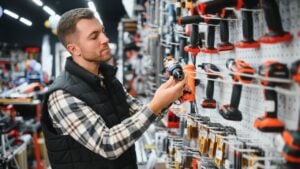 Portrait of young bearded man buying modern electric drill while standing in tool. Hardware construction site objects concept.