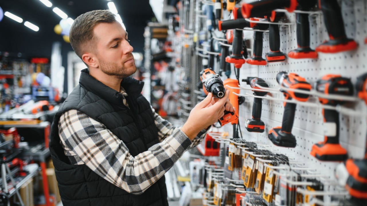 Portrait of young bearded man buying modern electric drill while standing in tool. Hardware construction site objects concept.
