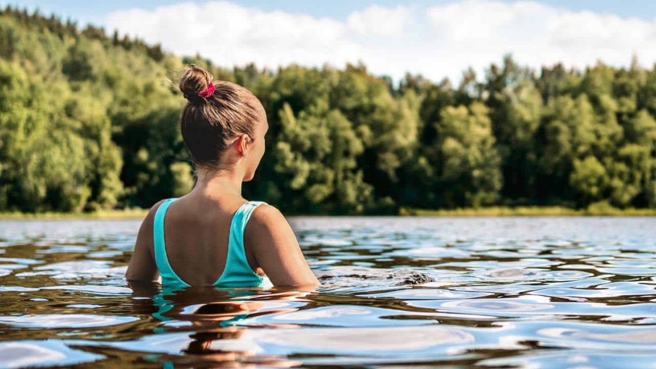 Swimming in lake in nature of Finland. Woman, water and forest. Summer at beach. Finnish bathing in river. Back view of person. Blue sky and beautiful green trees. Happy people relax in Scandinavia.