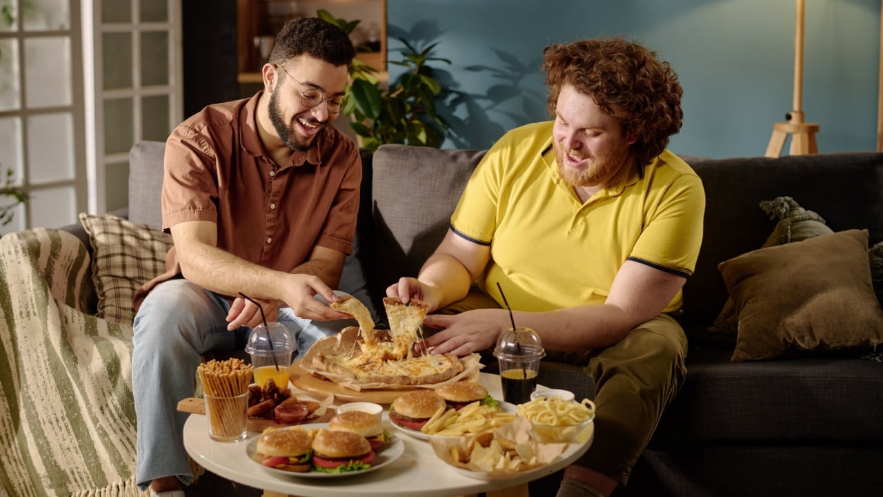 Two friends are laughing while enjoying a variety of snacks and fast food on a cozy sofa in a warmly-lit living room with colorful decor. Middle-aged men are having fun