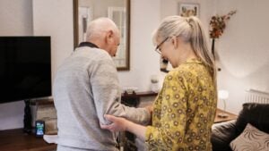 Love, help and senior couple in a living room with balance, support or walking in nursing home together. Mobility, care or elderly people in a lounge for stroke recovery, standing or healing activity