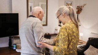 Love, help and senior couple in a living room with balance, support or walking in nursing home together. Mobility, care or elderly people in a lounge for stroke recovery, standing or healing activity