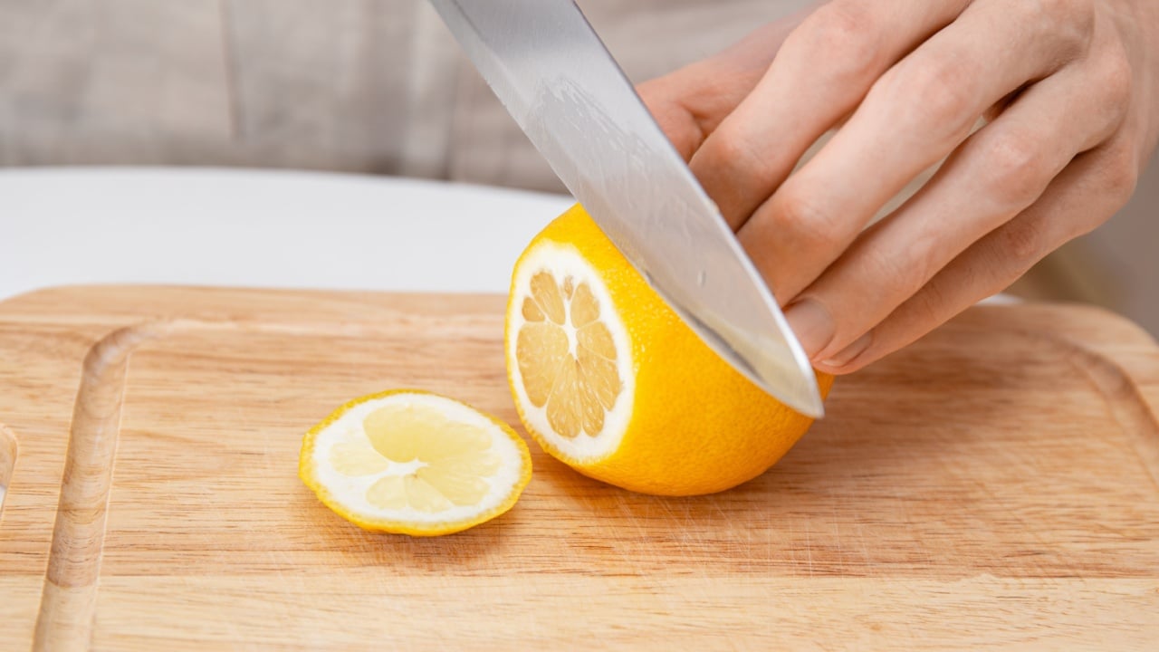 Close-up of a person slicing a fresh lemon with a kitchen knife on a wooden cutting board. Cooking prep, citrus