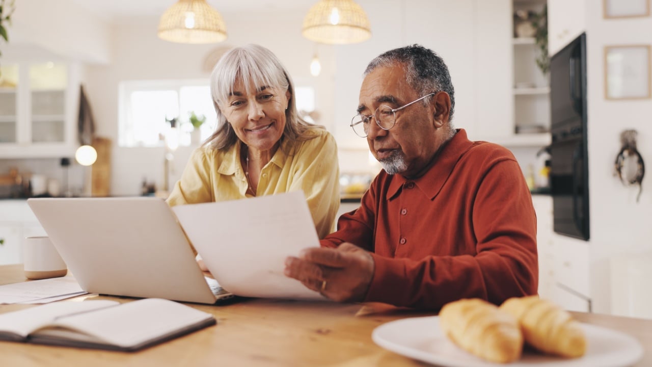 Elderly couple, documents and finance in home for results, laptop or smile with goal for pension. Interracial relationship, man and woman for investing, happy or profit for retirement income in house