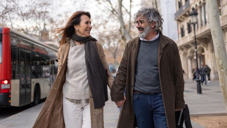 Happy senior couple holding hands and strolling through the bustling city center, enjoying their vacation together while wheeling their luggage along the vibrant streets