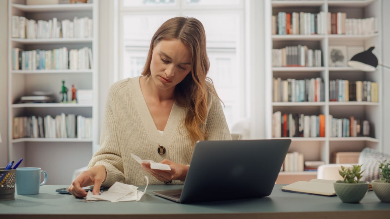 White Woman in Cream Cardigan Reviews Receipts at Her Laptop. Softly Lit Room With Bookshelves, Woman Does Taxes and BIlls, Organizing Financial Documents, Checks. Honest Taxpayer