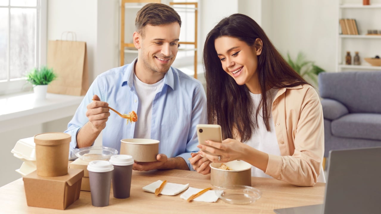 Happy young married couple sitting at table with laptop eating fresh fast food looking on mobile phone screen having a lunch together making online order. Take away food and delivery service concept.