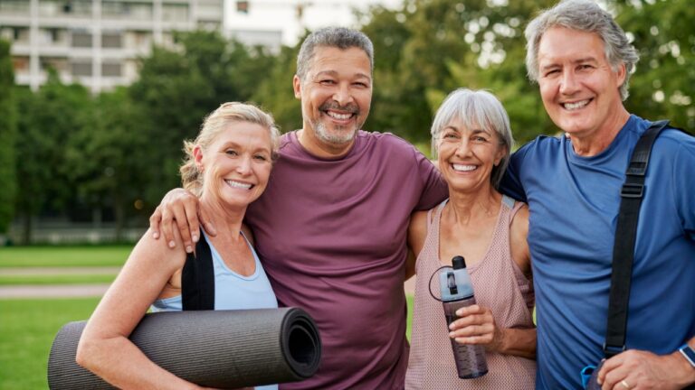 Four seniors smiling and looking at camera after exercising in the park. Multiethnic mature friends enjoying happy moments together with gym bags. Group hug and laughter after a fitness session.