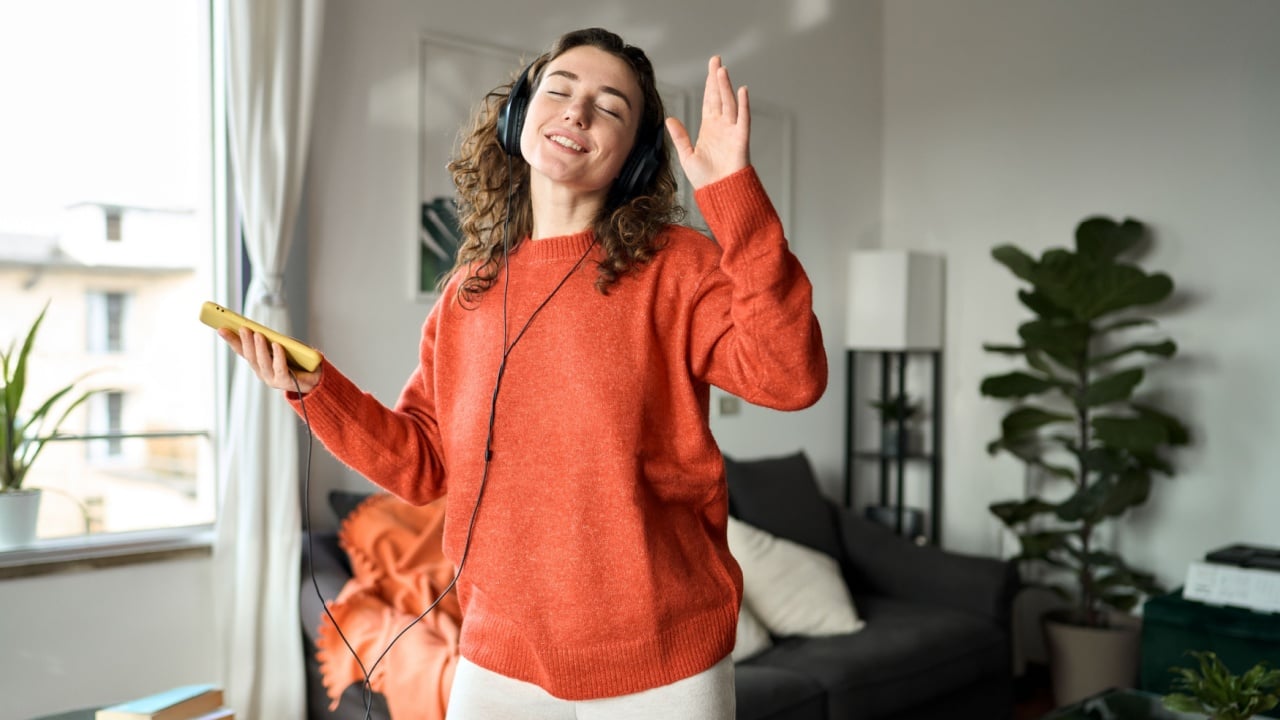 Candid photo of happy pretty young woman wearing headphones dancing at home. Cheerful girl listening music on mobile phone, singing song feeling relaxed standing in modern living room interior.