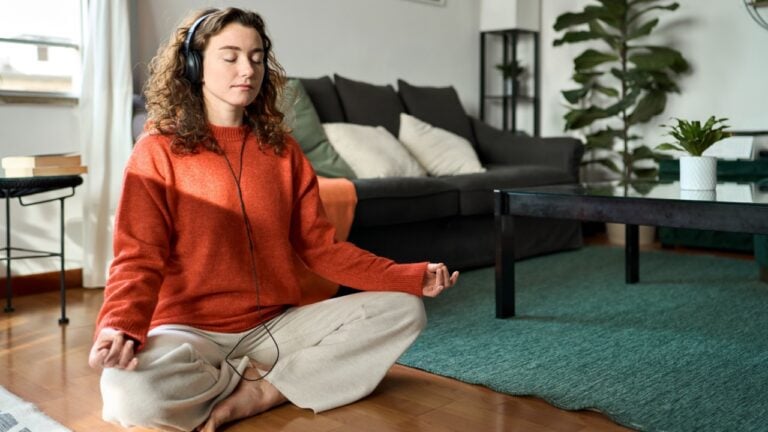 Mindful girl doing yoga meditation relaxing exercise for mental balance in apartment. Young pretty healthy woman wearing headphones listening calming music audio podcast sitting on floor at home.