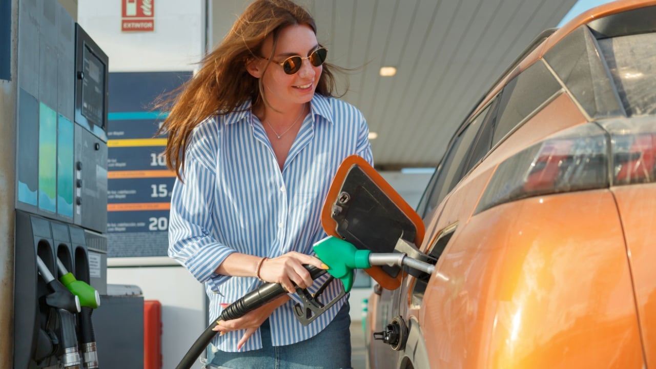 A woman in sunglasses fills her orange car with fuel at a gas station. The bright daylight highlights her casual summer outfit and cheerful demeanor.