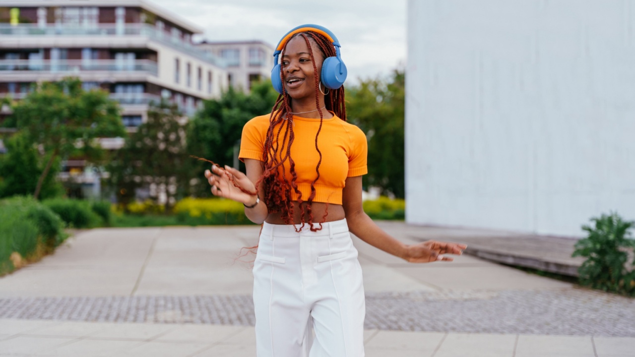 Black woman in orange crop top and white pants wearing headphones dances in city street. Urban background with buildings, greenery and street art. Full body, motion moment