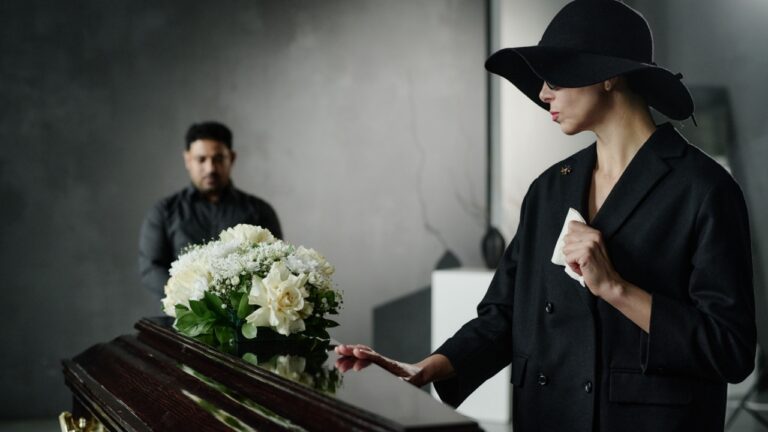 Caucasian woman wearing black hat standing beside coffin touching wood surface holding tissue while man standing in background near floral arrangement during funeral
