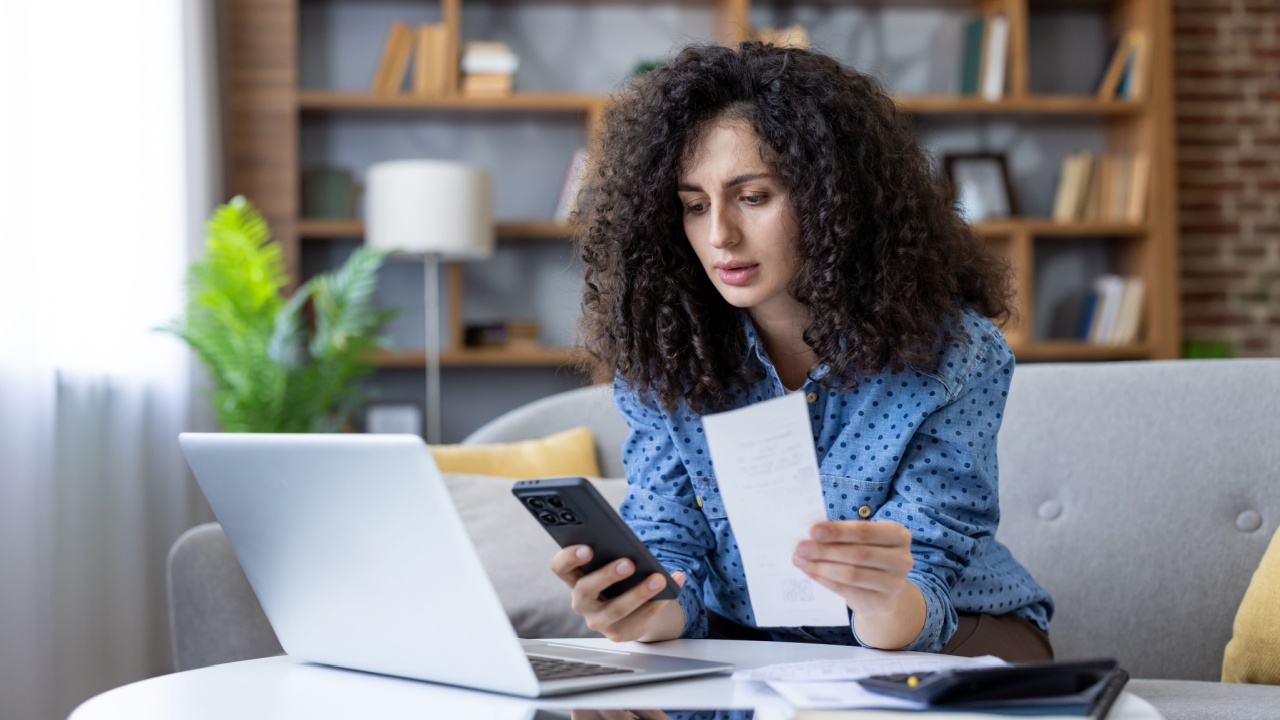 Woman reviewing household finances on laptop and phone, calculating bills and receipts in living room while managing budget, online payments and personal expenses