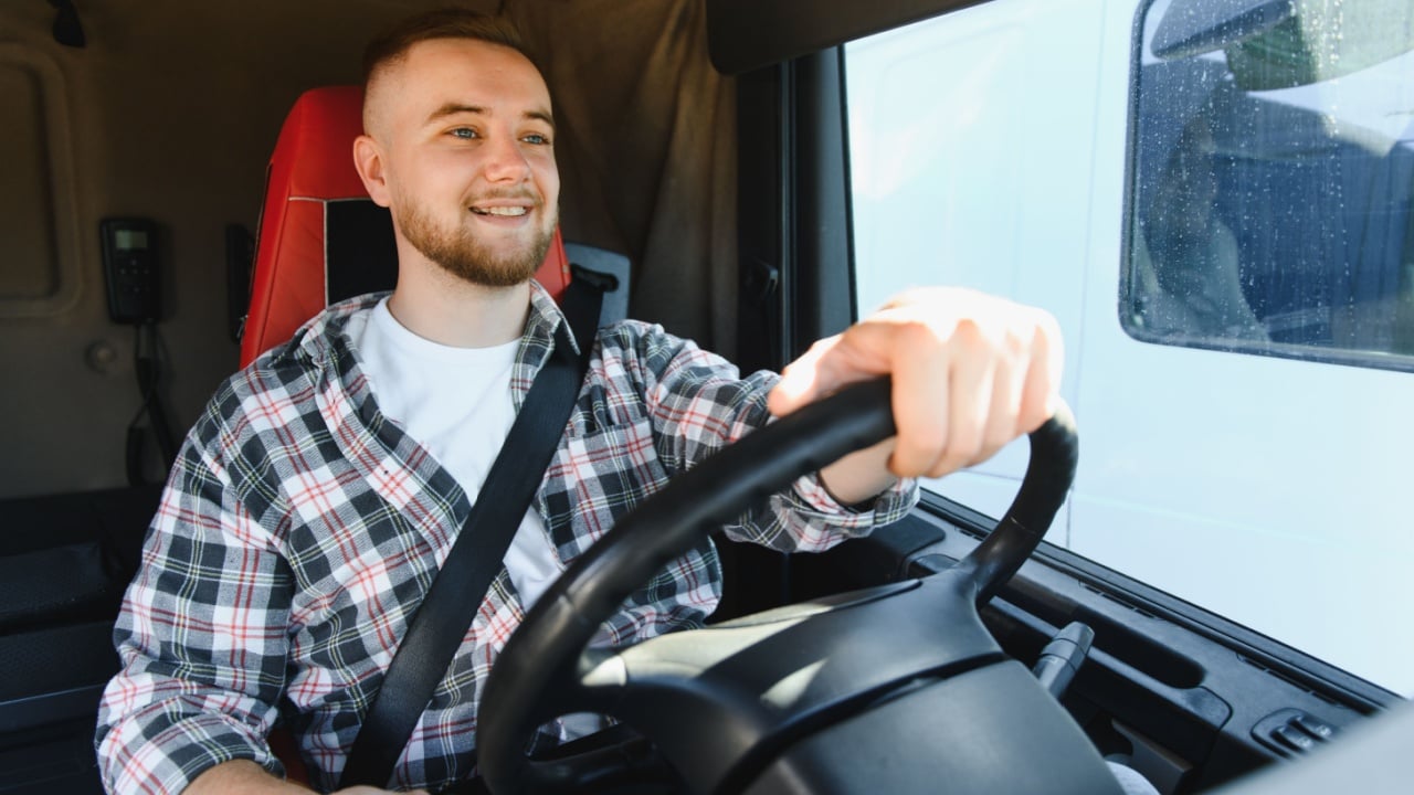 Young male truck driver smiling, wearing a seatbelt and holding a steering wheel, driving a semi truck