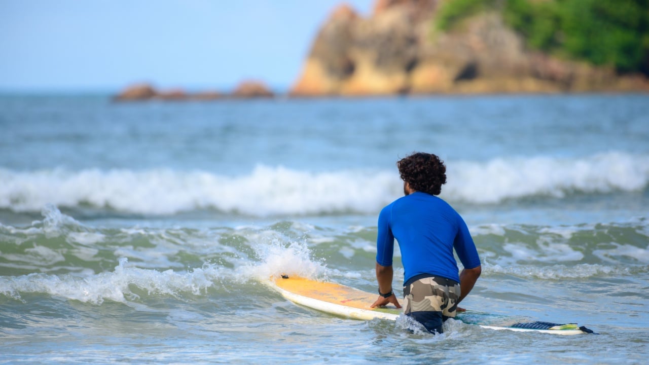 Weligama, Sri Lanka - 05 29 2023: Surfer boy with curly hair wearing a blue rash guard and camouflage shorts kneels on his surfboard, preparing to paddle into the gentle wave