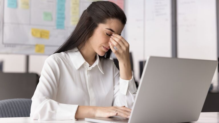 Too much screen time. Tired fatigued young female office worker sit at desk by notebook with closed eyes rub nose bridge suffer from migraine eye strain tension headache stressed with mental burnout