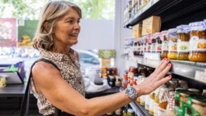 Senior woman smiling and reaching for a jar on a supermarket shelf, enjoying the shopping experience and considering her product choices in a grocery store