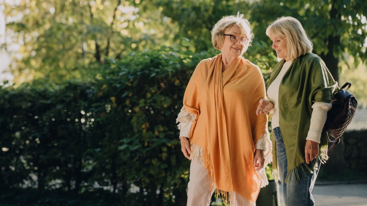 Two elderly female friends walk arm in arm through a sunny park, enjoying conversation and companionship in their retirement