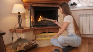 Woman in casual clothing sitting comfortably on the floor, adding a piece of firewood to a burning fireplace, creating a warm and inviting atmosphere during cold weather at home