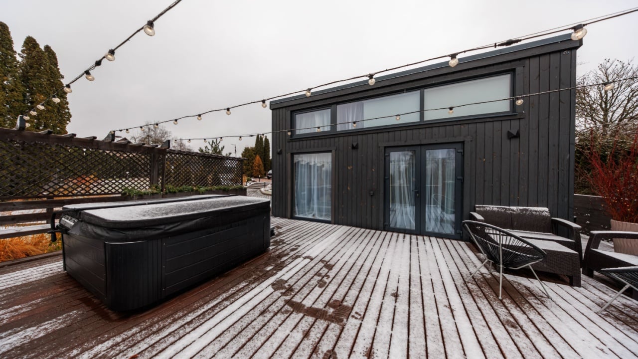 A dark tiny house with tall clerestory windows stands on a snowy deck, string lights hang overhead, footprints lead to a hot tub and lounge chairs in overcast daylight.
