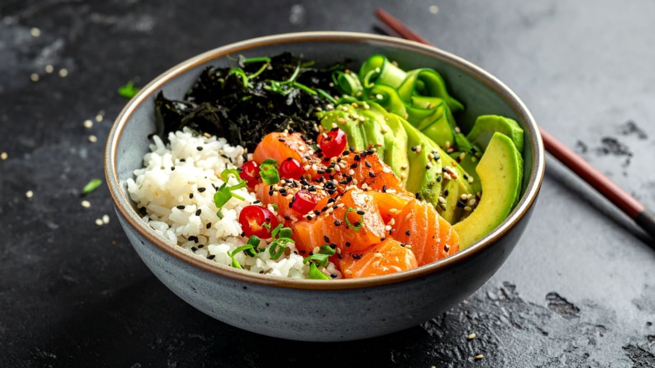 A poke bowl with salmon, rice, avocado, seaweed, and other toppings is shown in a bowl with chopsticks.
