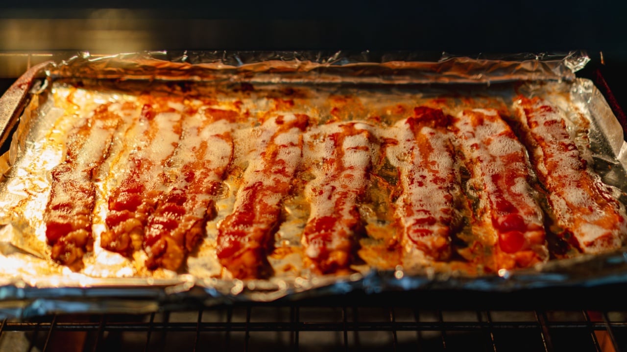 Crispy bacon strips sizzling on a foil-lined baking tray in an oven.