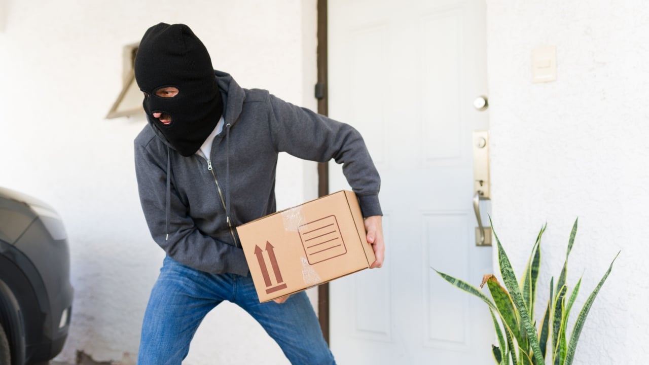 Thief wearing a black balaclava and grey hoodie quickly running away with a stolen cardboard box from a house porch