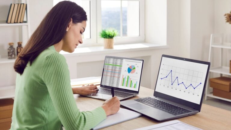 Analyst woman reviews charts and graphs on laptops. She reviews metrics at a bright office desk, notes trends in a notebook, and interprets performance. Clear concept of data analytics.