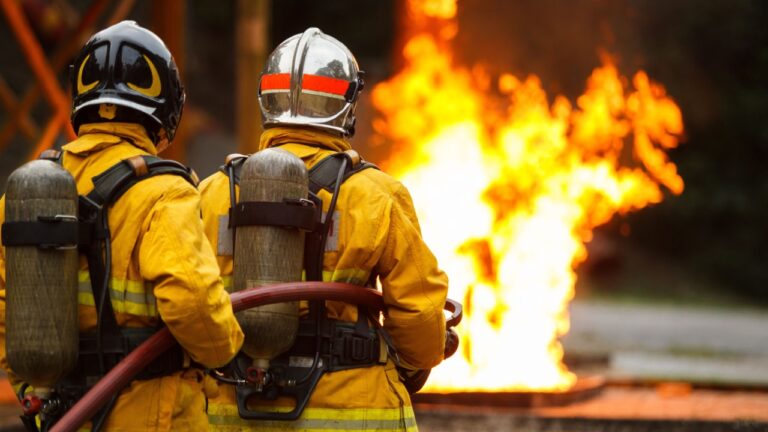 firefighters team wearing full protective gear operate fire hose to control large flames during active fire. Emergency response, teamwork, fire suppression, industrial safety, and disaster management.