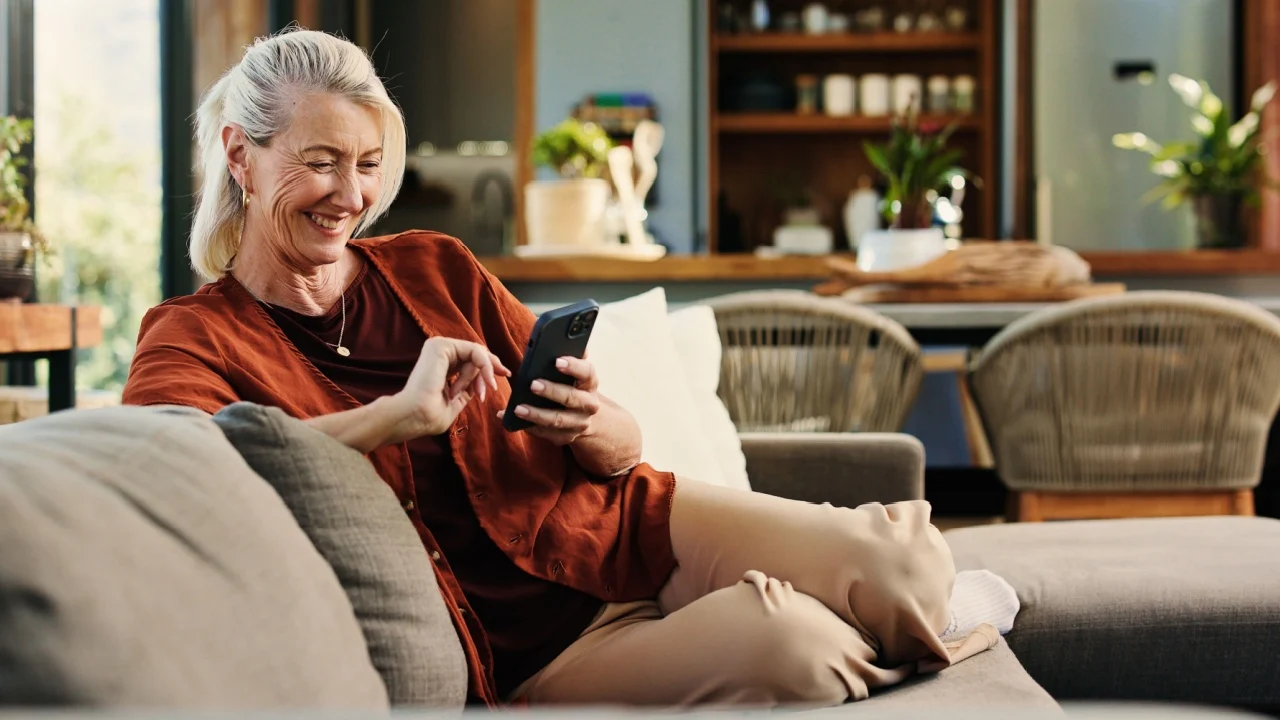 Old woman, phone and laugh on sofa with texting, notification and funny chat in living room at house. Person, smartphone and happy with mobile app, contact and social network on couch at apartment