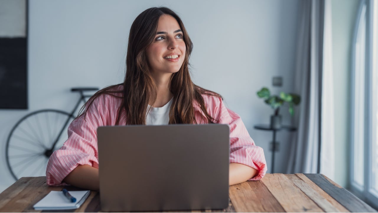 A dreamy young woman sits at her desk with a laptop, looking into the distance while taking a break from work, reflecting on ideas and visualizing plans in a calm home environment