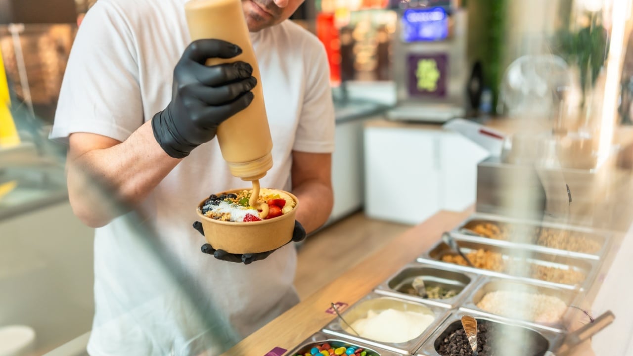 Barista wearing black gloves preparing a customized acai bowl, adding a creamy sauce over fresh fruits, granola, and other various toppings in a paper cup in a modern cafe setting