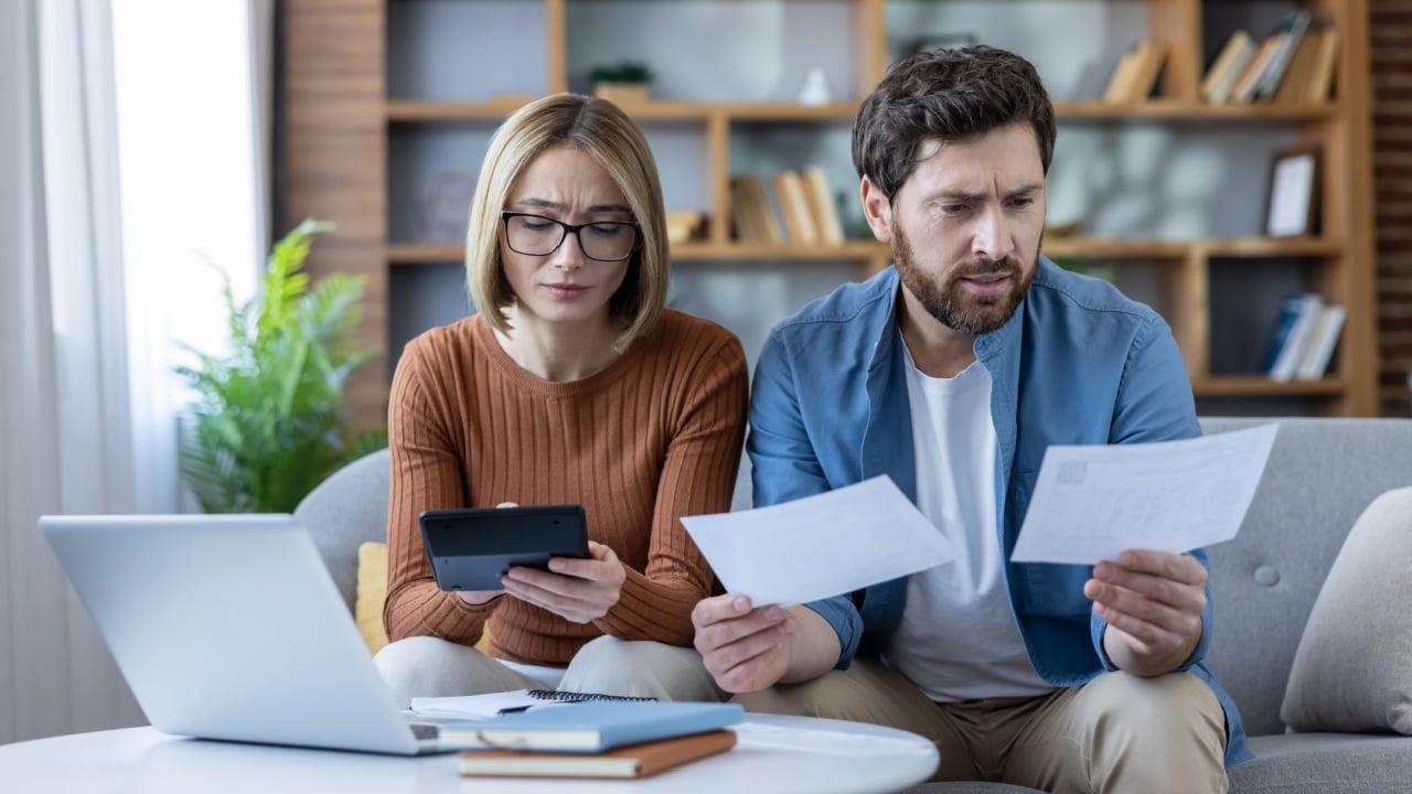 Stressed couple analyzing financial bills and expenses, using a calculator and laptop to manage debt and household budget, feeling worried about their money situation