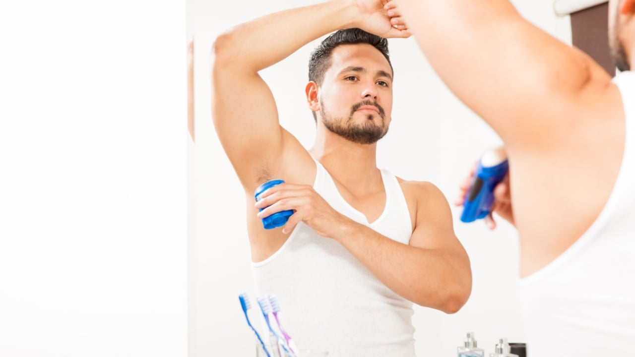 Good looking young man with a beard putting on some deodorant in front of a mirror in a bathroom