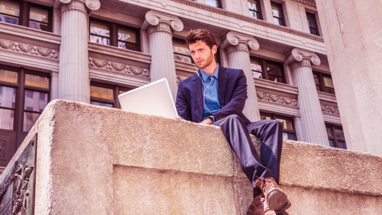 European Business Man working on street in New York. Dressing in blue suit, brown leather boot shoes, wearing ear phone, young guy with beard, reading, typing on laptop computer