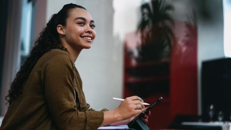Smiling Latina entrepreneur working remotely from trendy café,using digital tablet and stylus for efficient project management.