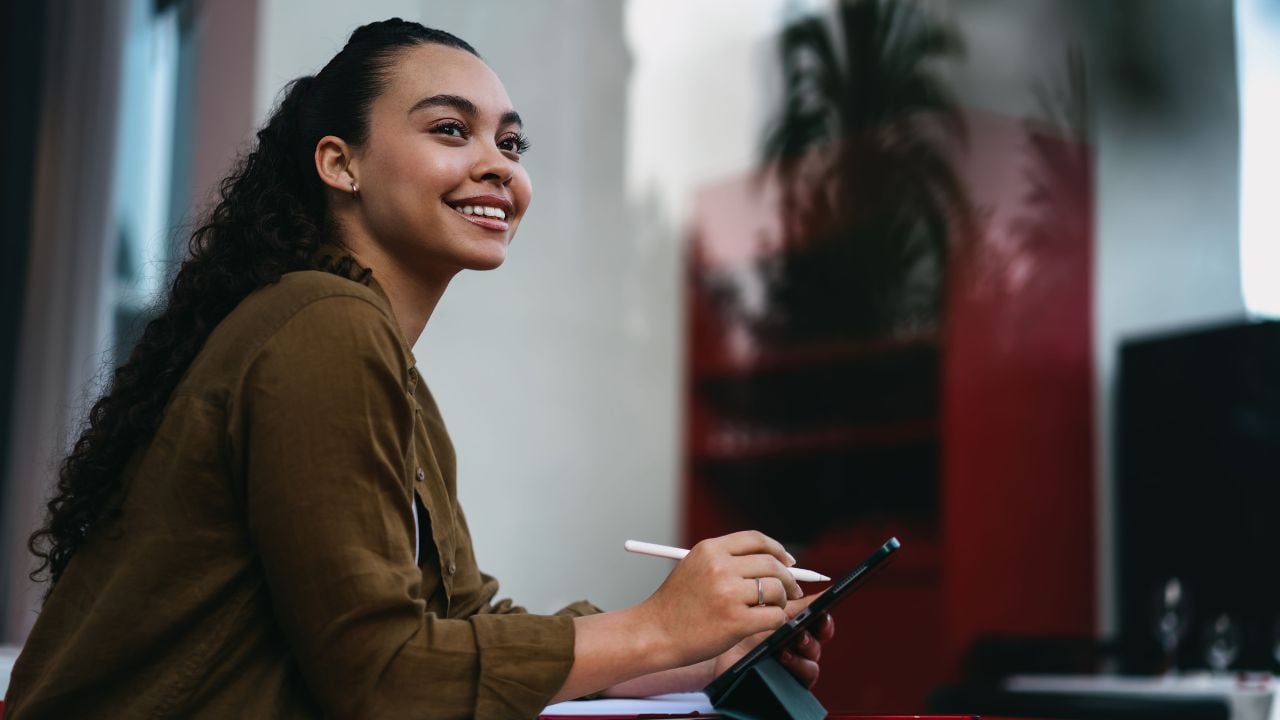 Smiling Latina entrepreneur working remotely from trendy café,using digital tablet and stylus for efficient project management.