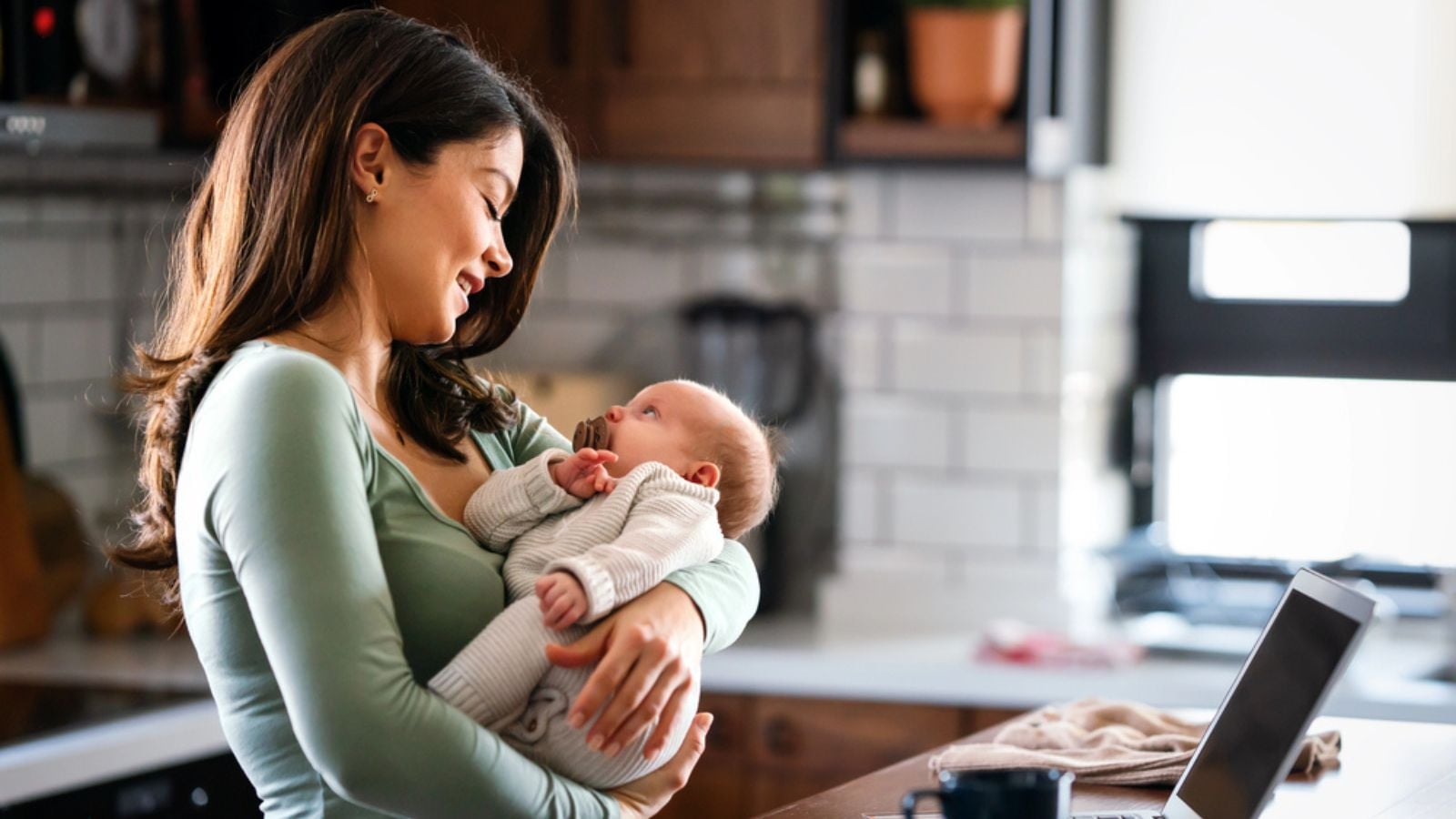 stay at home mom working remotely on laptop holding baby infant